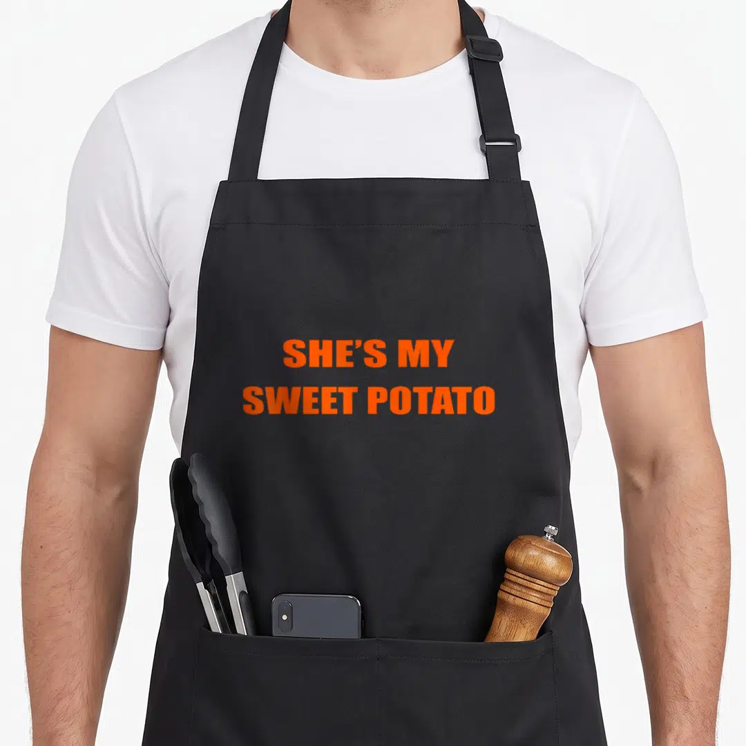Goal potato apron - Orange text reading She's My Sweet Potato stacked in two lines — shown on a black cotton canvas apron against a white background