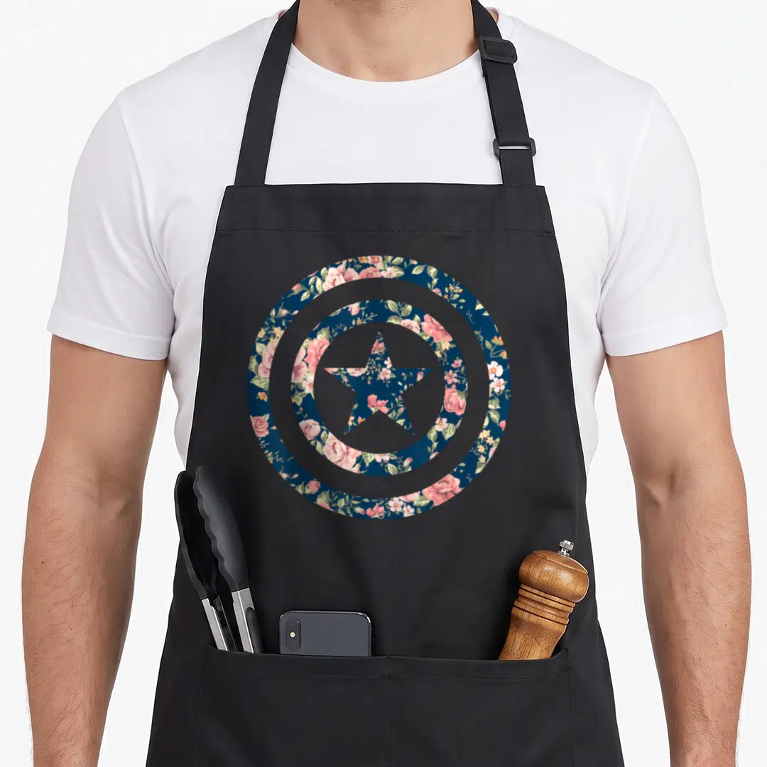 Icon floral apron - Circular shield and star filled with pink, green, and dark blue floral pattern — shown on a black cotton canvas apron against a white background