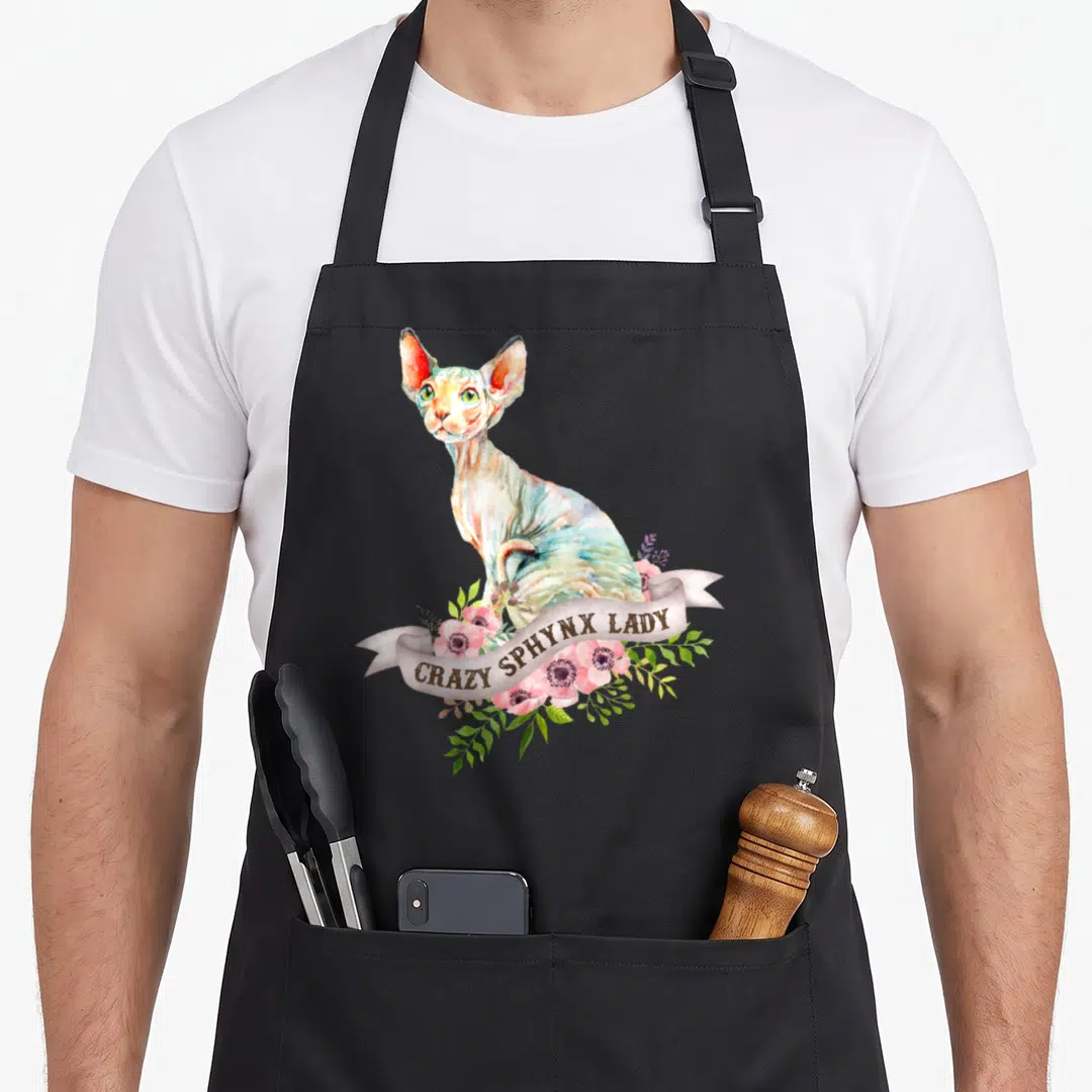 Sphynx crazy cat lady apron - Watercolor Sphynx cat sitting above a ribbon with pink flowers and green leaves reading — shown on a black cotton canvas apron against a white background