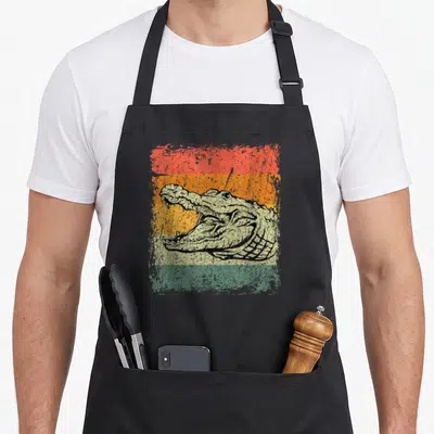Cute crocodile alligator apron - Distressed crocodile head with open mouth over red orange yellow green beige stripes — shown on a black cotton canvas apron against a white background