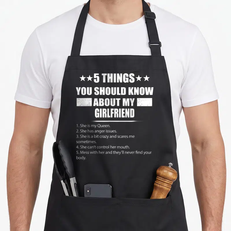 Girlfriend novelty apron - White distressed text listing 5 facts about a girlfriend including anger issues and Queen — shown on a black cotton canvas apron against a white background