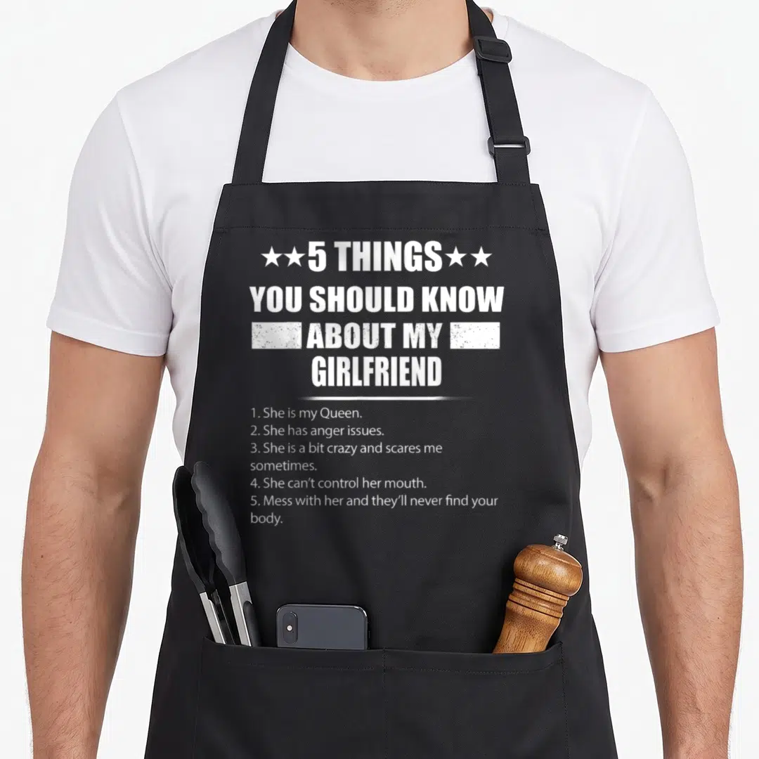 Girlfriend novelty apron - White distressed text listing 5 facts about a girlfriend including anger issues and Queen — shown on a black cotton canvas apron against a white background