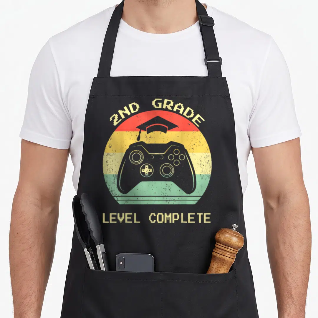Graduation 2nd apron - Retro game controller and cap inside striped circle with 2ND GRADE LEVEL COMPLETE text — shown on a black cotton canvas apron against a white background