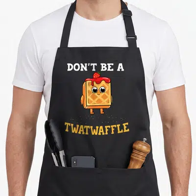 Twatwaffle maker apron - Cartoon waffle with red syrup and strawberry under text Don't Be A Twatwaffle — shown on a black cotton canvas apron against a white background