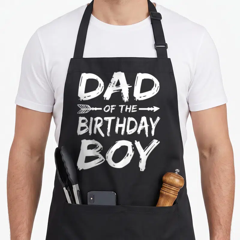 Cool birthday family cooking aprons - DAD OF THE BIRTHDAY BOY distressed white text with arrow graphic — shown on a black cotton canvas apron against a white background