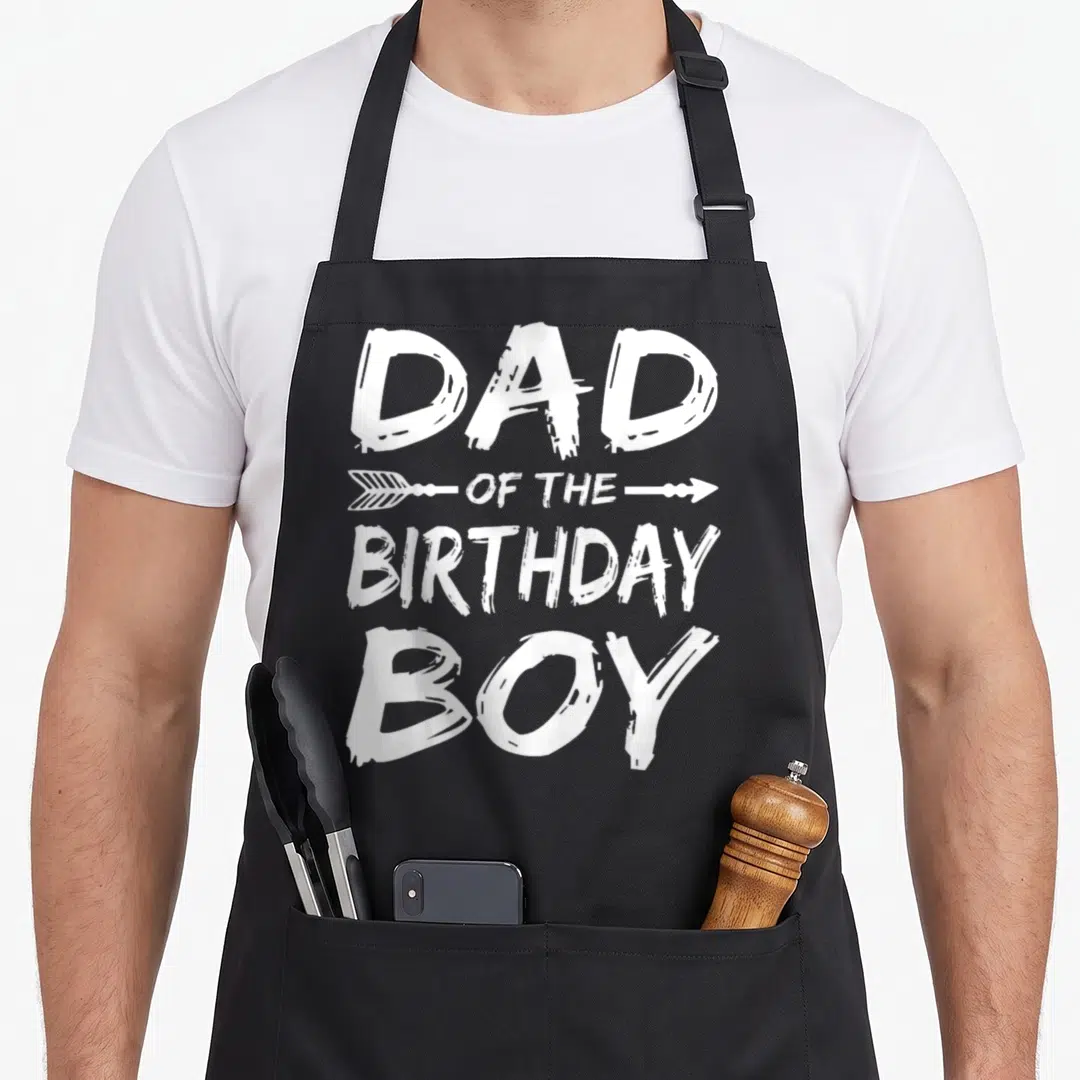 Cool birthday family cooking aprons - DAD OF THE BIRTHDAY BOY distressed white text with arrow graphic — shown on a black cotton canvas apron against a white background