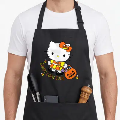 Cutie halloween apron - Hello Kitty holding pumpkin bucket with candy corn and arched text in orange yellow white black — shown on a black cotton canvas apron against a white background