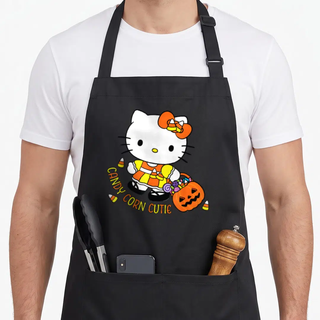 Cutie halloween apron - Hello Kitty holding pumpkin bucket with candy corn and arched text in orange yellow white black — shown on a black cotton canvas apron against a white background