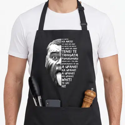 Lyric new zealand apron - Black and white photo of a man's face with tongue out next to stacked white haka lyrics — shown on a black cotton canvas apron against a white background