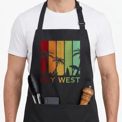 Key west florida apron - Retro graphic with red orange yellow green stripes, grey palm trees, surfboard, and KEY WEST — shown on a black cotton canvas apron against a white background