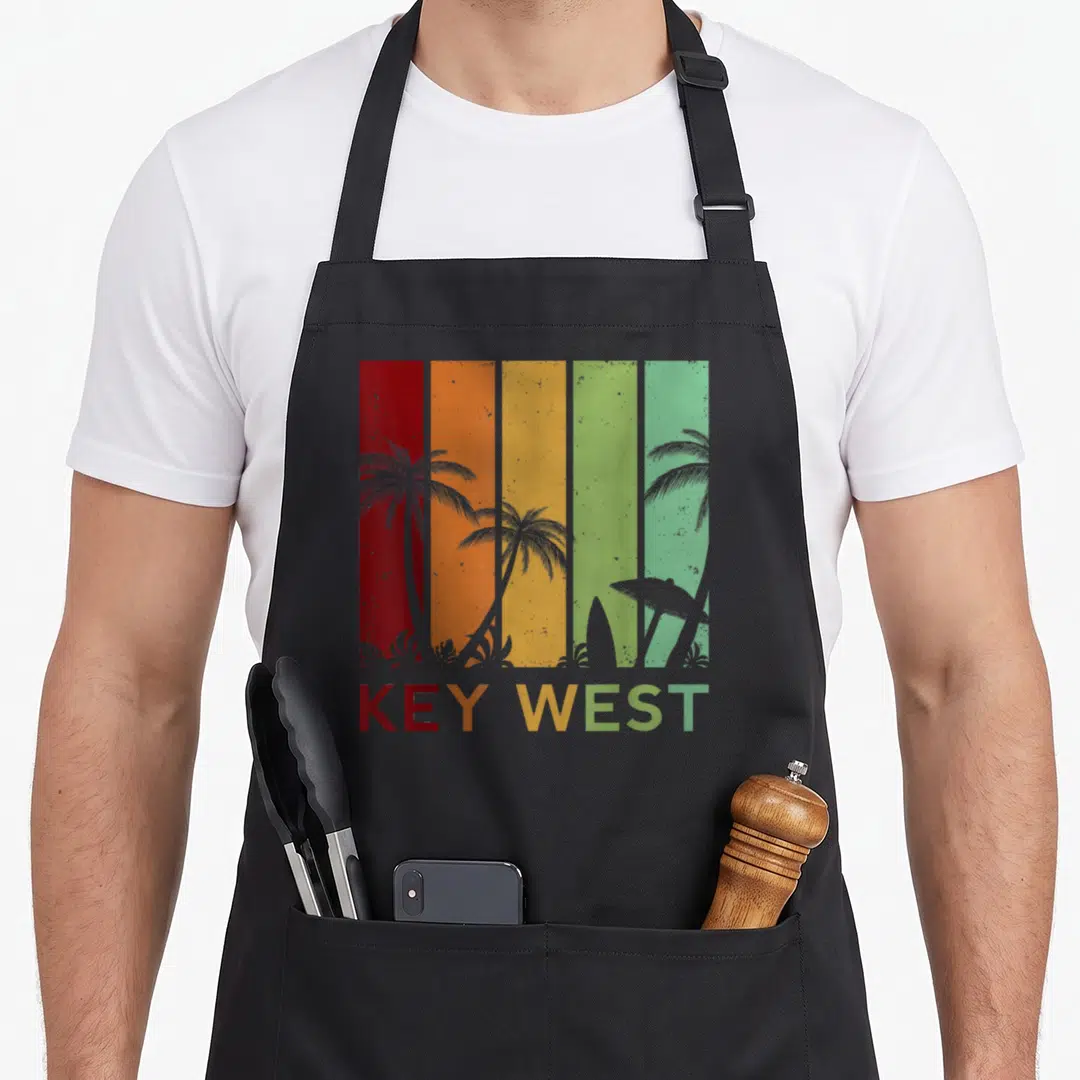 Key west florida apron - Retro graphic with red orange yellow green stripes, grey palm trees, surfboard, and KEY WEST — shown on a black cotton canvas apron against a white background