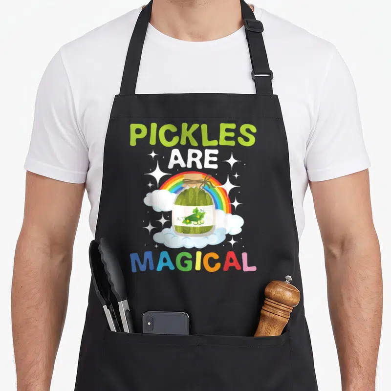 Magical vegetable apron - Pickle jar on clouds with rainbow and text PICKLES ARE MAGICAL — shown on a black cotton canvas apron against a white background