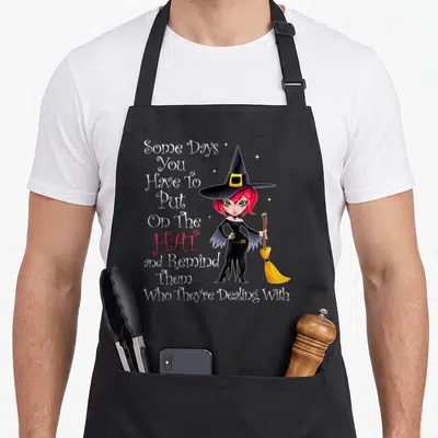 Hat broom halloween witch apron - Cartoon witch holding broom next to stacked text Some Days You Have To Put On The Hat — shown on a black cotton canvas apron against a white background