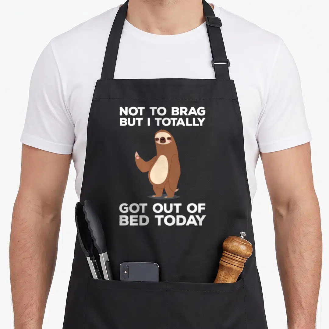 Bed sloth apron - Cartoon brown sloth standing between white text reading NOT TO BRAG BUT I TOTALLY GOT OUT OF BED TODAY — shown on a black cotton canvas apron against a white background