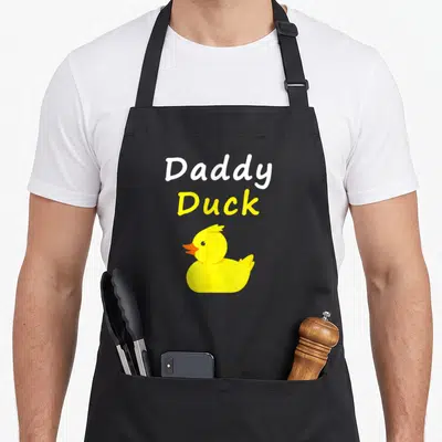 Daddy cotton duck apron - Stacked Daddy Duck text above a yellow cartoon rubber duck with an orange beak — shown on a black cotton canvas apron against a white background