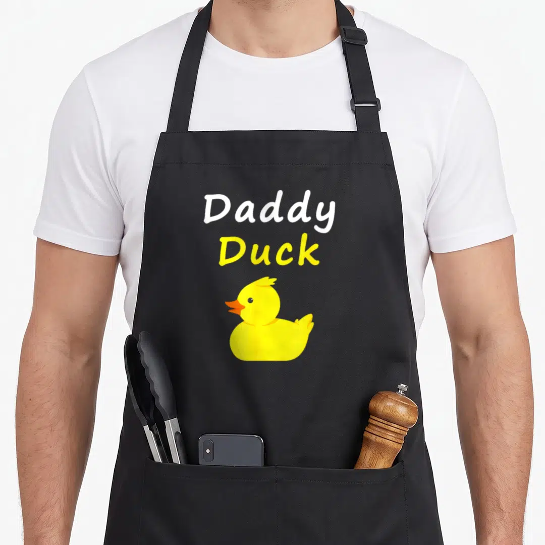 Daddy cotton duck apron - Stacked Daddy Duck text above a yellow cartoon rubber duck with an orange beak — shown on a black cotton canvas apron against a white background
