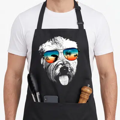 Wheaten coated apron - Shaggy terrier face wearing aviator sunglasses reflecting a beach scene with sailboat and palm — shown on a black cotton canvas apron against a white background