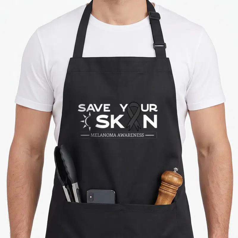 Save melanoma awareness apron - White SAVE YOUR SKIN text with sun icon and black ribbon replacing the I in SKIN above — shown on a black cotton canvas apron against a white background