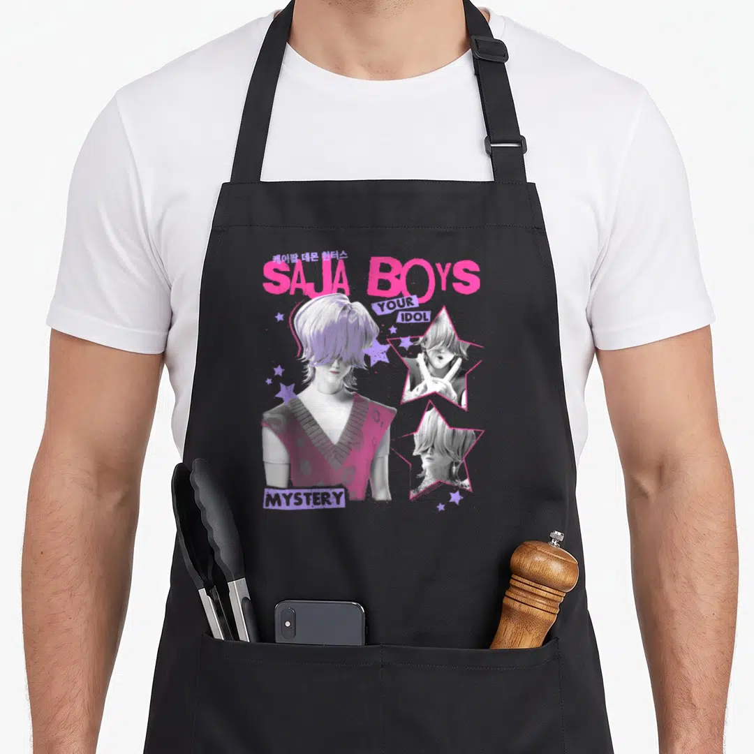 Saja mystery apron - SAJA BOYS text with man portrait and two star-framed images in pink purple grey white — shown on a black cotton canvas apron against a white background