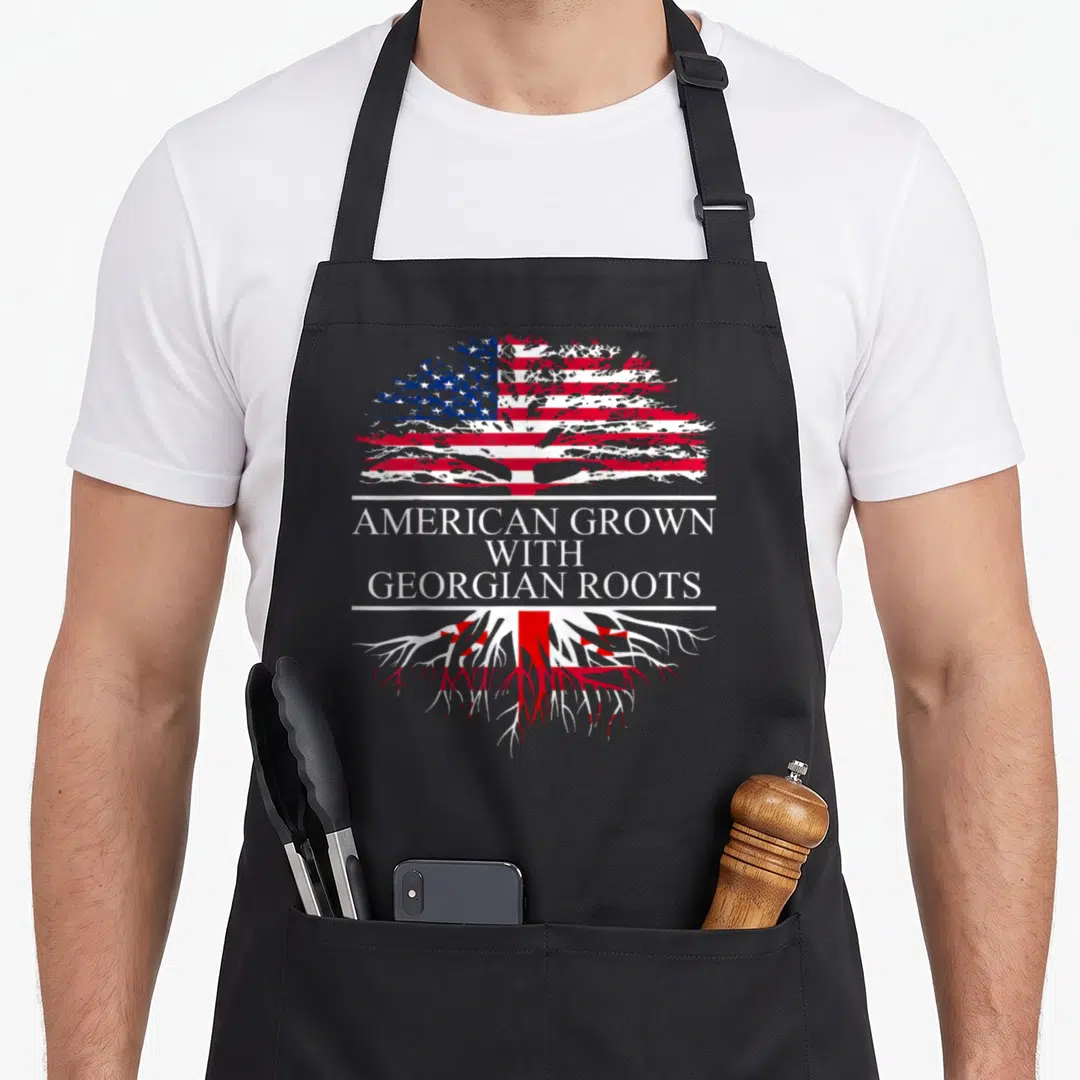 Georgian root american flag apron - Tree with American flag canopy and Georgian flag roots framing stacked white text — shown on a black cotton canvas apron against a white background