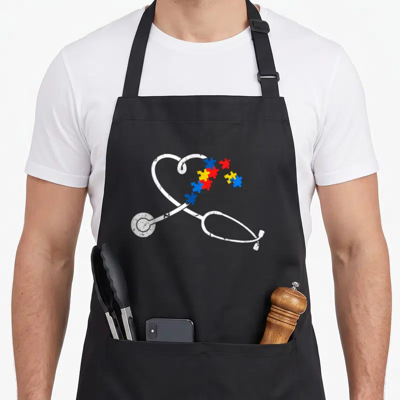 Autism nurse apron - White stethoscope heart shape filled with blue red and yellow puzzle pieces — shown on a black cotton canvas apron against a white background