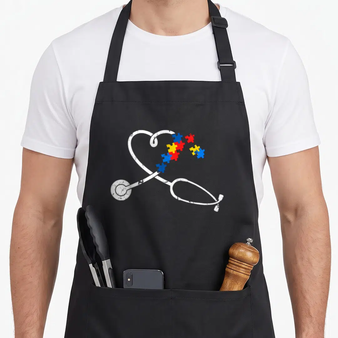 Autism nurse apron - White stethoscope heart shape filled with blue red and yellow puzzle pieces — shown on a black cotton canvas apron against a white background