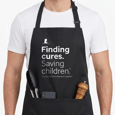 Jude st apron - White silhouette child and text Finding cures Saving children on black background — shown on a black cotton canvas apron against a white background