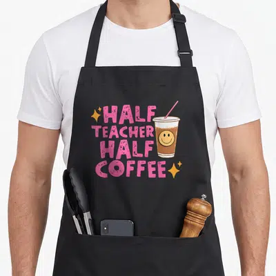 Coffee teacher half apron - Stacked pink text reading HALF TEACHER HALF COFFEE beside a smiling iced coffee cup — shown on a black cotton canvas apron against a white background