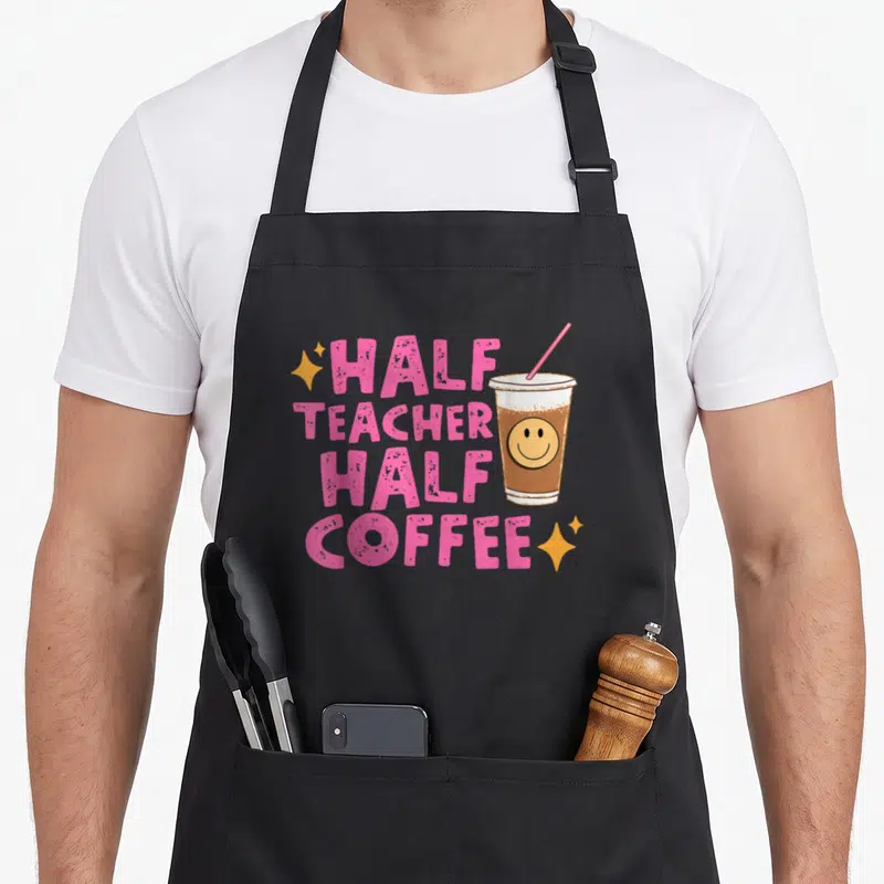 Coffee teacher half apron - Stacked pink text reading HALF TEACHER HALF COFFEE beside a smiling iced coffee cup — shown on a black cotton canvas apron against a white background