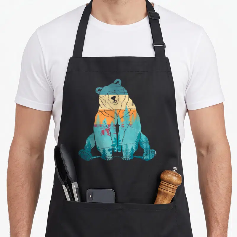 Animal forest bear apron - Retro bear silhouette filled with horizontal blue teal orange yellow grey gradient and pine — shown on a black cotton canvas apron against a white background
