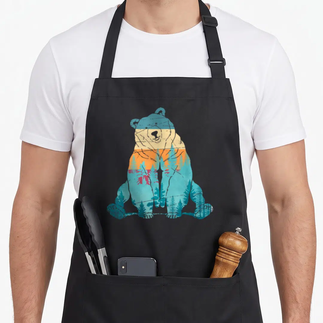 Animal forest bear apron - Retro bear silhouette filled with horizontal blue teal orange yellow grey gradient and pine — shown on a black cotton canvas apron against a white background