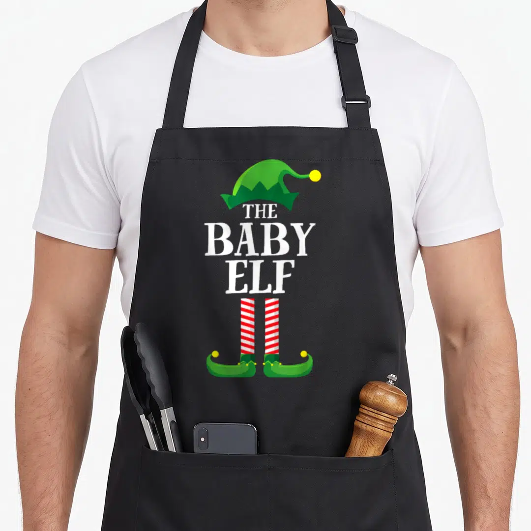 Baby personalized family aprons - Stacked text The Baby Elf with green hat, red striped legs, and curled green shoes — shown on a black cotton canvas apron against a white background