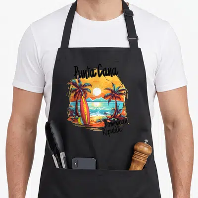 Punta family aprons - Punta Cana arched text, summer surfboard, palm trees, sunset, Dominican Republic label — shown on a black cotton canvas apron against a white background
