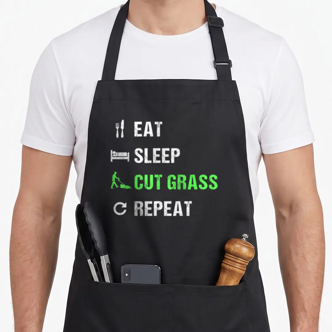 Eat landscaper apron - Stacked EAT SLEEP CUT GRASS REPEAT text with fork knife bed mower repeat icons in white and green — shown on a black cotton canvas apron against a white background