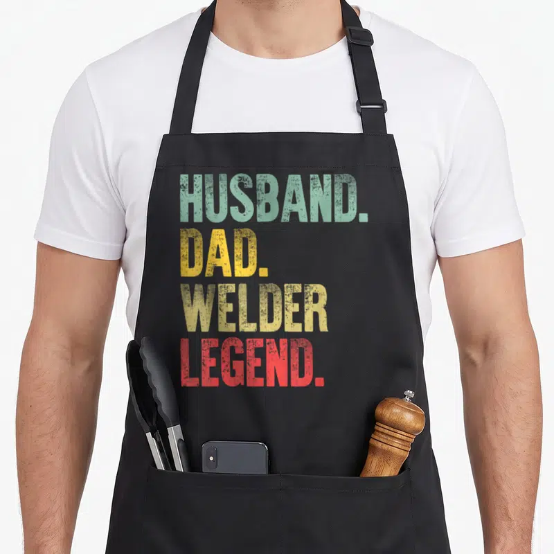 Welder husband apron - Stacked HUSBAND. DAD. WELDER. LEGEND. text in green, yellow, and red distressed font — shown on a black cotton canvas apron against a white background