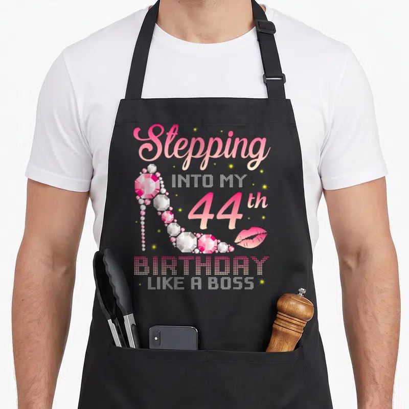 44th bbq boss apron - Pink diamond high heel shoe surrounded by Stepping Into My 44th Birthday Like A Boss text and lips — shown on a black cotton canvas apron against a white background