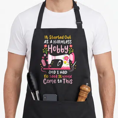 Sewing quilting quilt apron - Cartoon sewing machine with pink flowers and text about harmless hobby — shown on a black cotton canvas apron against a white background
