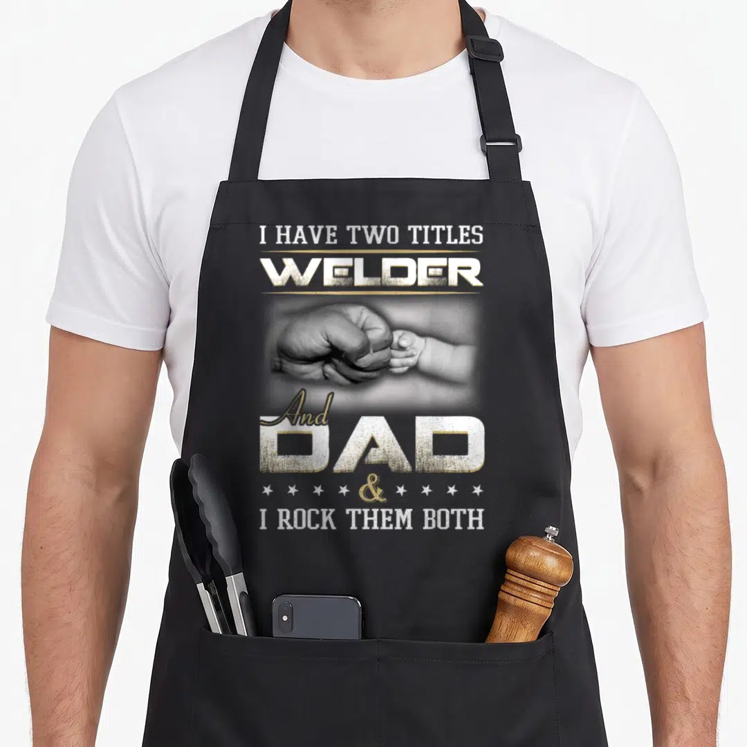 Welding daddy apron - I HAVE TWO TITLES WELDER And DAD I ROCK THEM BOTH with adult and baby hands fist bumping — shown on a black cotton canvas apron against a white background