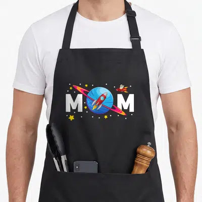 Funny outer birthday apron - MOM text split by blue planet red rocket astronaut yellow stars white dots — shown on a black cotton canvas apron against a white background