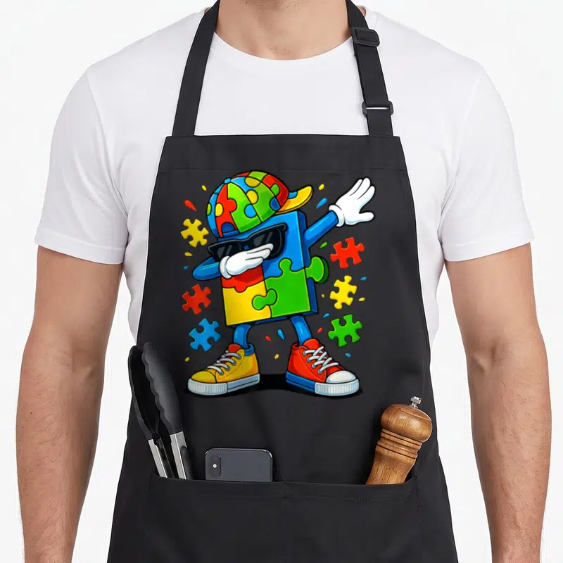 Dabbing autism awareness apron - Cartoon blue puzzle character dabbing in sunglasses and cap with scattered red yellow — shown on a black cotton canvas apron against a white background
