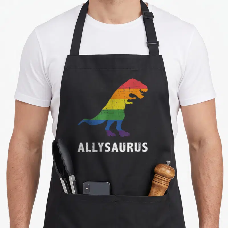 Allysaurus dinosaur apron - Rainbow striped T-Rex silhouette above white ALLYSAURUS text on grey background — shown on a black cotton canvas apron against a white background