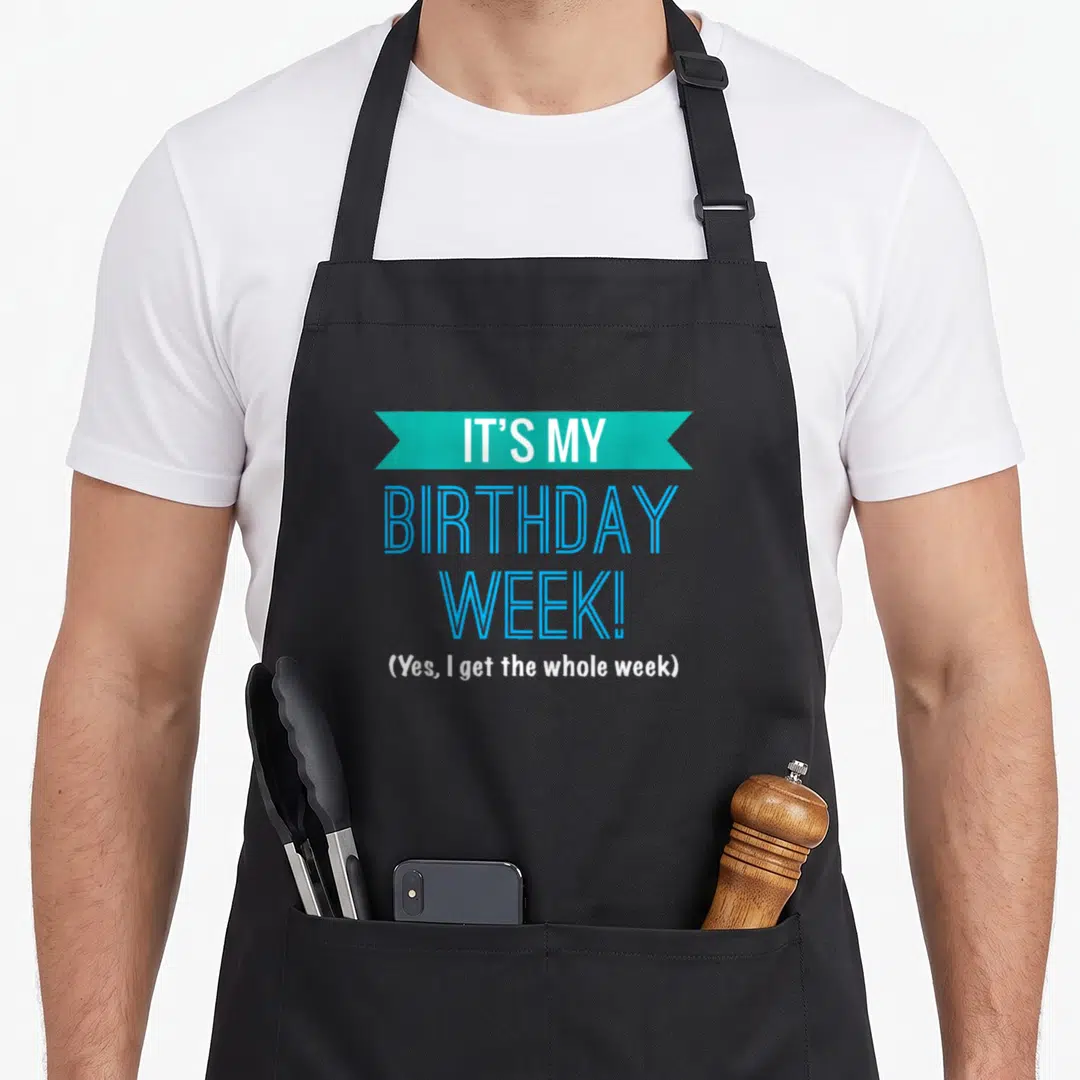 Retro week birthday apron - Teal ribbon banner with white text IT'S MY BIRTHDAY WEEK! above blue stacked lettering — shown on a black cotton canvas apron against a white background