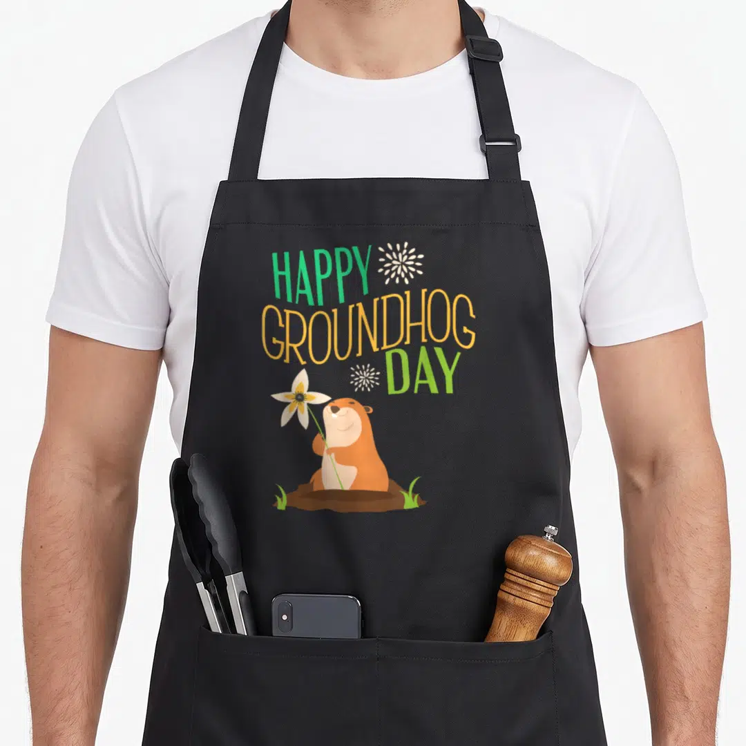 Ground groundhog apron - Cartoon groundhog holding a white flower under stacked green and orange text with white bursts — shown on a black cotton canvas apron against a white background
