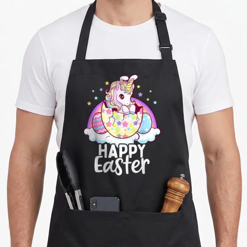 Unicorn easter egg apron - Cartoon unicorn hatching from star-patterned egg under rainbow with happy easter text — shown on a black cotton canvas apron against a white background