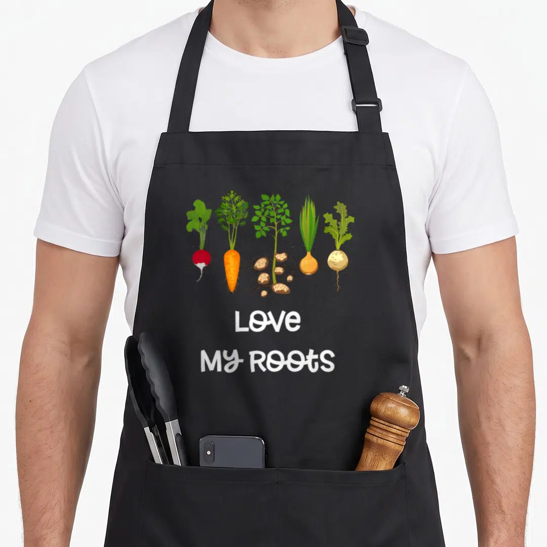 Root vegetable apron - Five cartoon root vegetables with Love My Roots text below — shown on a black cotton canvas apron against a white background