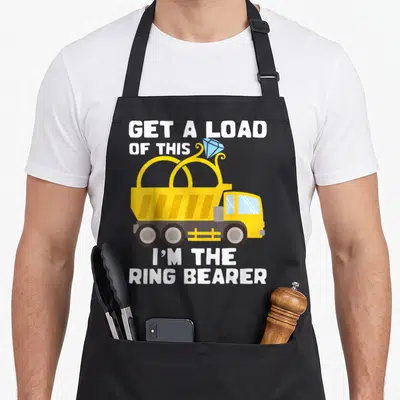 Bearer ring apron - Yellow dump truck carrying blue diamond ring with white text GET A LOAD OF THIS I'M THE RING BEARER — shown on a black cotton canvas apron against a white background