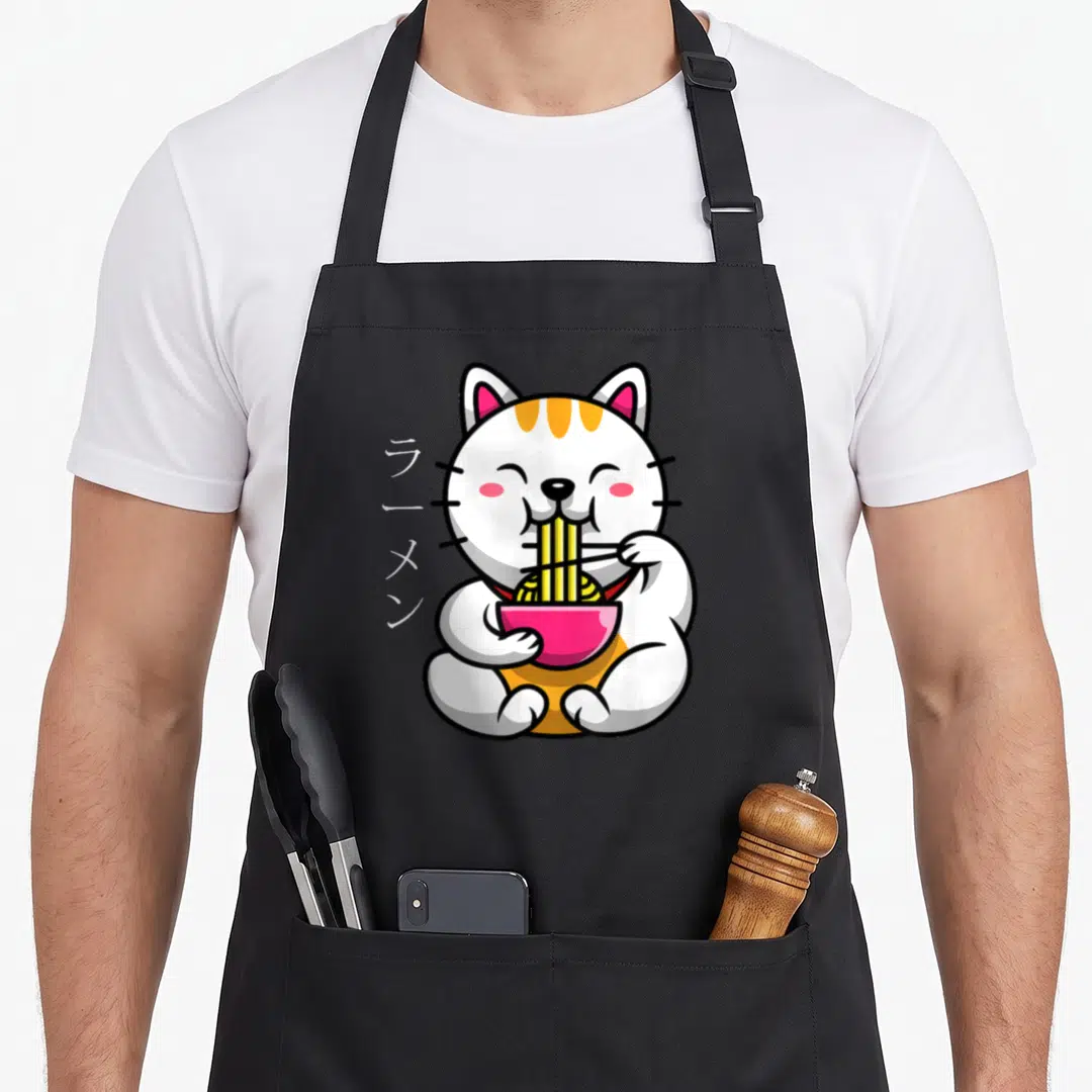 Ramen bowl cat apron - White cat with orange stripes eating yellow noodles from pink bowl next to vertical Japanese text — shown on a black cotton canvas apron against a white background