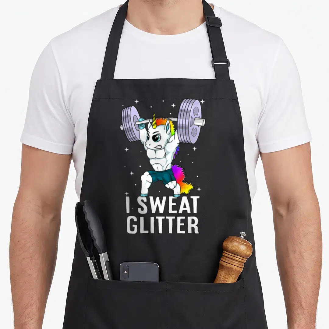 Sweat unicorn apron - Muscular white unicorn lifting purple barbell with I SWEAT GLITTER text and rainbow mane — shown on a black cotton canvas apron against a white background
