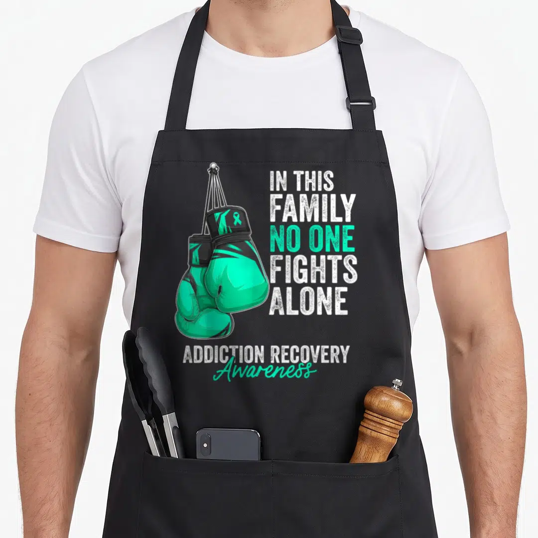 Addiction ribbon apron - Two teal boxing gloves hang beside IN THIS FAMILY NO ONE FIGHTS ALONE text with ADDICTION — shown on a black cotton canvas apron against a white background