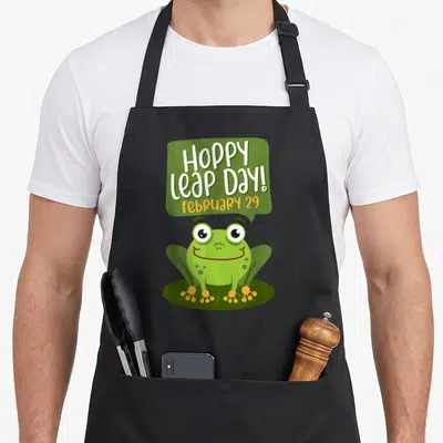 Hoppy frog aprons - Cartoon green frog on lily pad with Hoppy Leap Day text in speech bubble — shown on a black cotton canvas apron against a white background